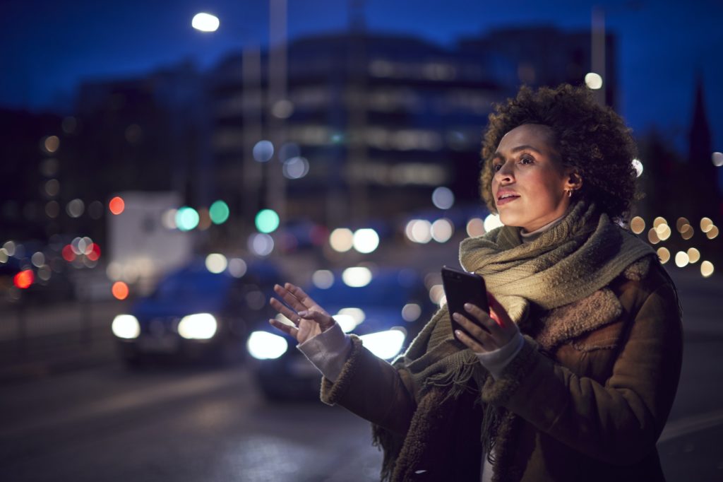 Woman On City Street At Night Ordering Taxi Using Mobile Phone App